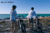 Discounted Blutron Sale Series Electric Bikes - Two individuals resting these promotional electric bicycles on a coastal gravel shoreline while enjoying the ocean view