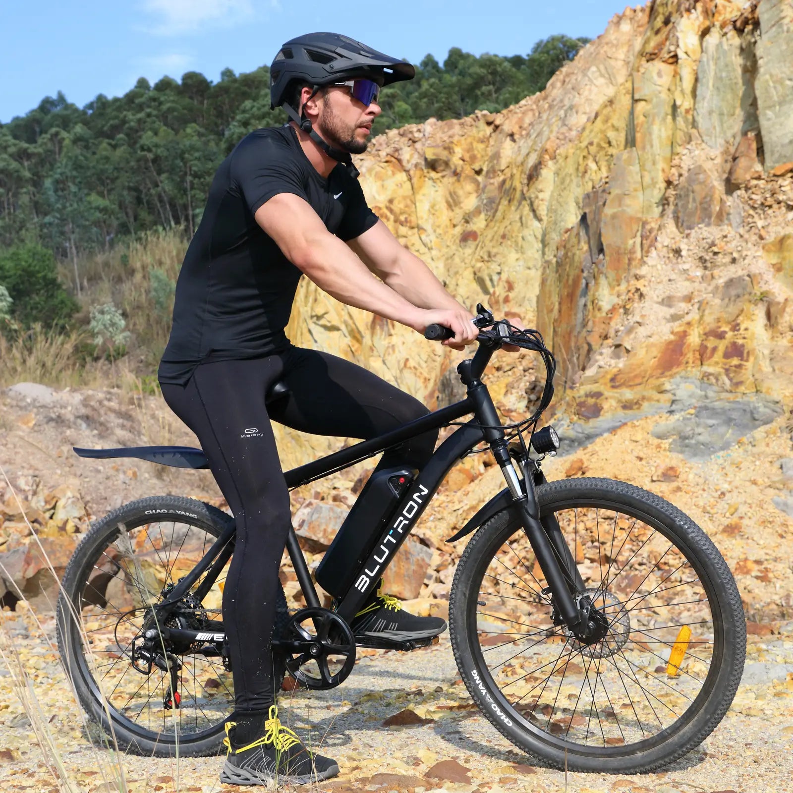 BLUTRON EB550 off-road electric mountain bike: Rider in athletic gear navigating gravel terrain, with the bike featuring a detachable BLUTRON-branded battery — set against a rocky, wooded outdoor backdrop