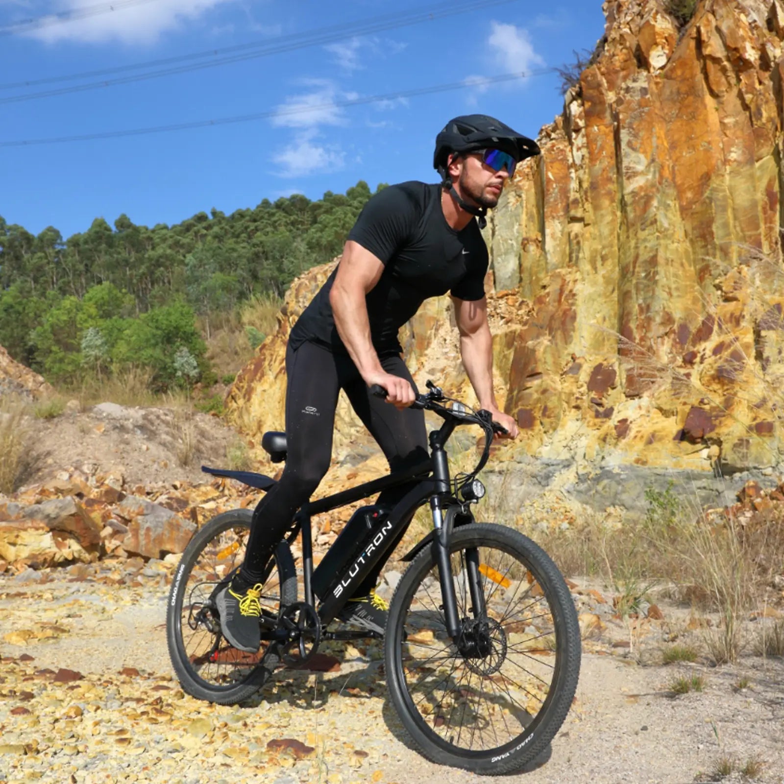 BLUTRON EB550 off-road ebike: Rider in athletic gear navigating rocky gravel terrain, featuring a detachable BLUTRON-branded battery — set against a rugged outdoor backdrop of cliffs and vegetation