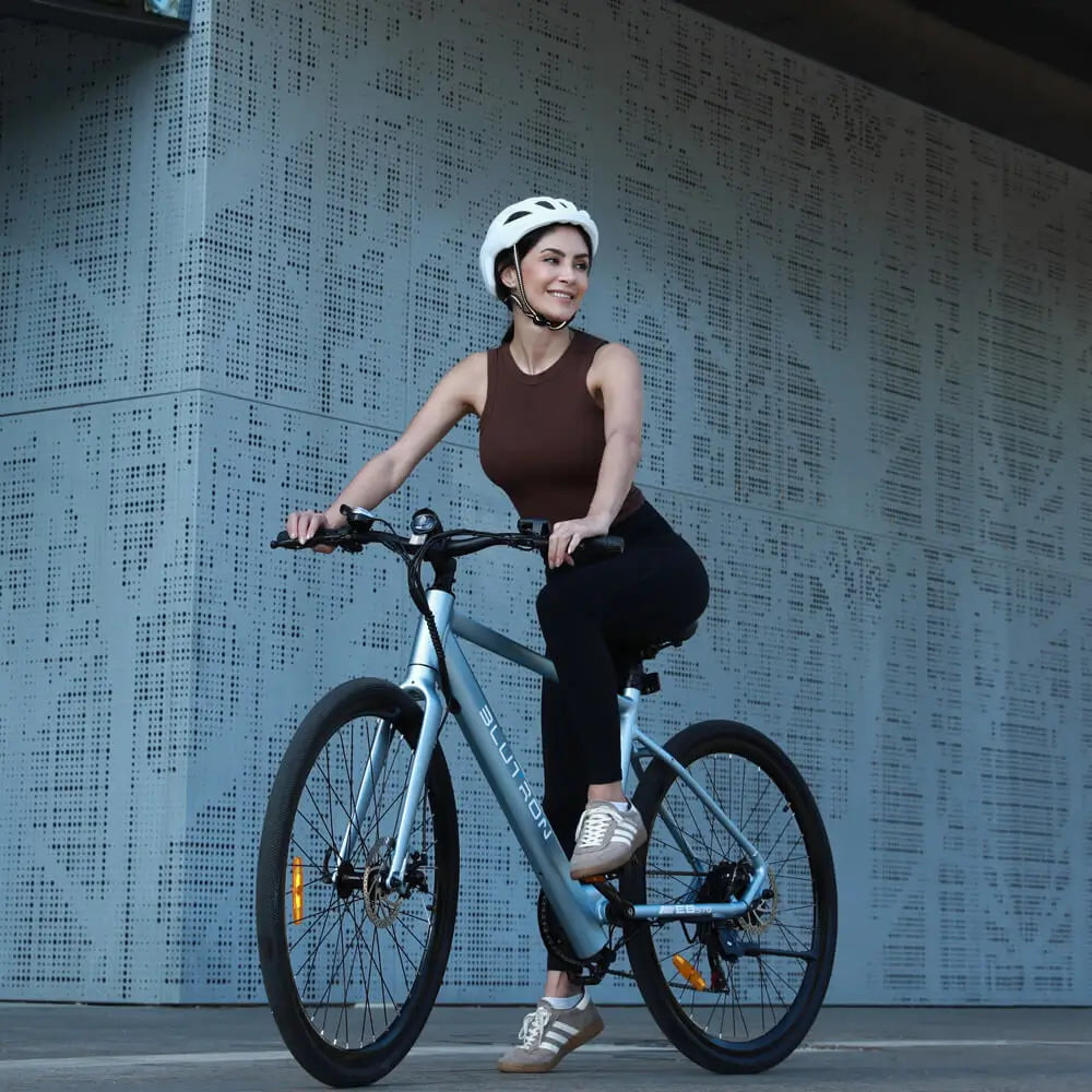 A helmeted woman rides the Blutron EB570—one of the best value commuter ebikes—by an urban building. It features a sleek frame and comfortable seating, ideal for affordable, smooth daily city commutes.