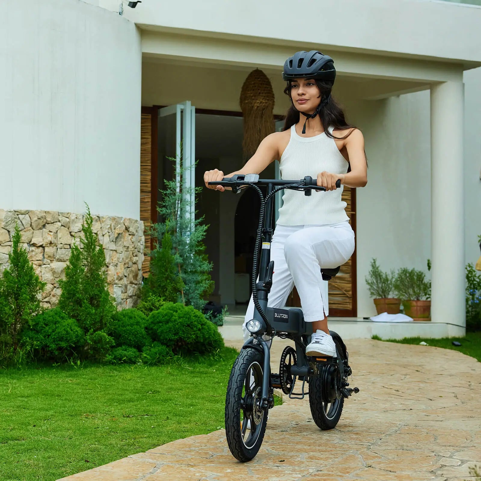 Helmeted woman riding a BLUTRON long range foldable ebike on a stone path outside a home (greenery backdrop) — the compact bike features a built-in LED headlight