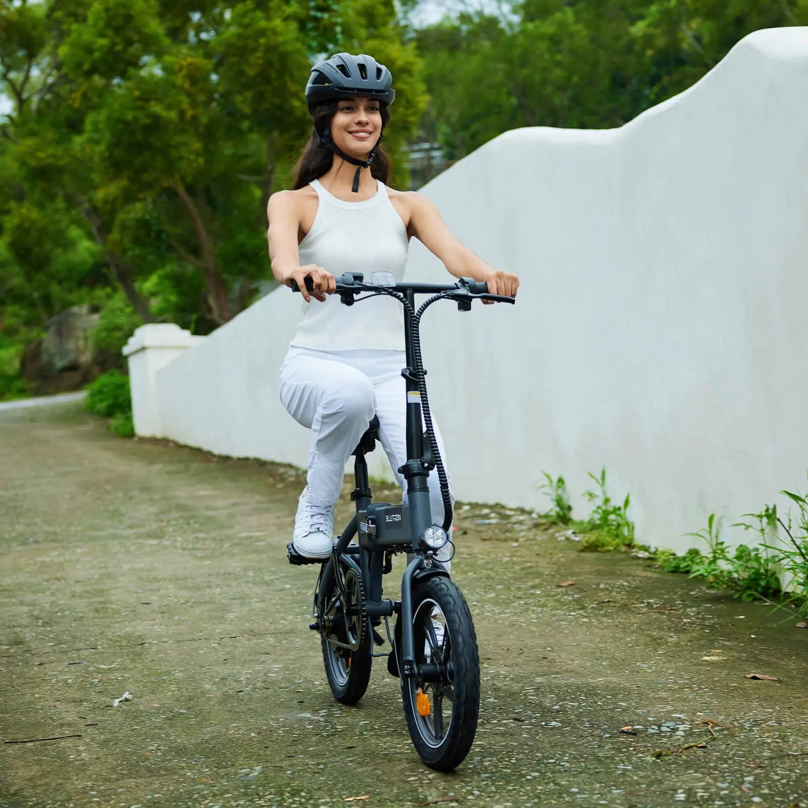 Helmeted woman riding a BLUTRON mid drive folding ebike along a paved path (flanked by a white wall and greenery) — the compact bike features a built-in LED headlight