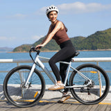 Helmeted woman rides the Blutron electric commuter ebike along a coastal boardwalk (sea, hills backdrop). It has disc brakes and a sleek frame—ideal for smooth daily commutes and leisure rides.