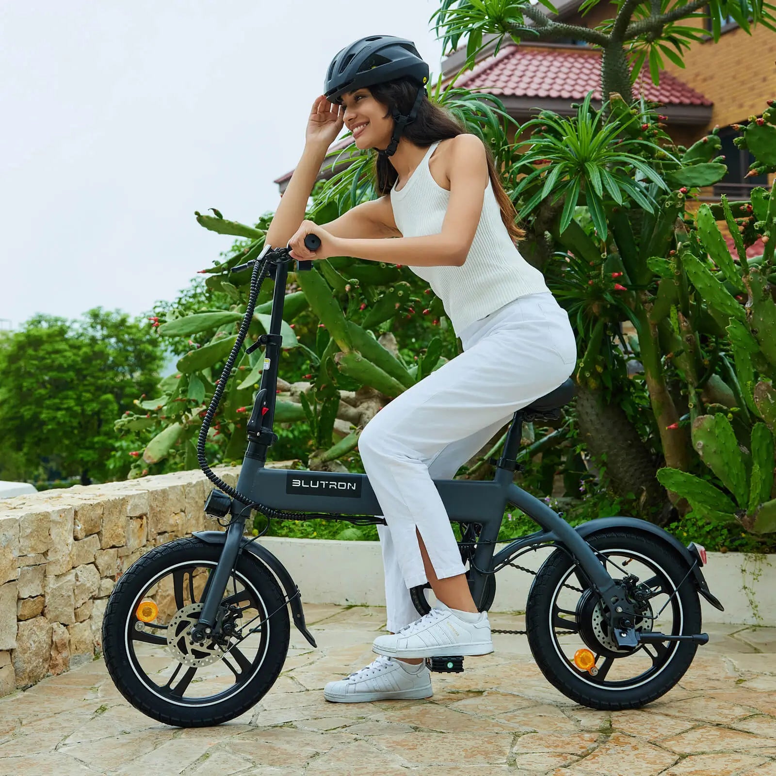 Helmeted woman riding a BLUTRON ebike (among top 10 folding ebikes) on a stone path — flanked by cacti and a stone wall, the bike features front/rear disc brakes and a sleek, compact frame