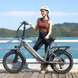 Woman with a white helmet standing beside a Blutron lightweight folding ebike: Silver-gray model featuring "BLUTRON" logo, rear cargo rack, wide tires, front/rear disc brakes, positioned on a coastal boardwalk with blue waters and green hills in the sunny background.