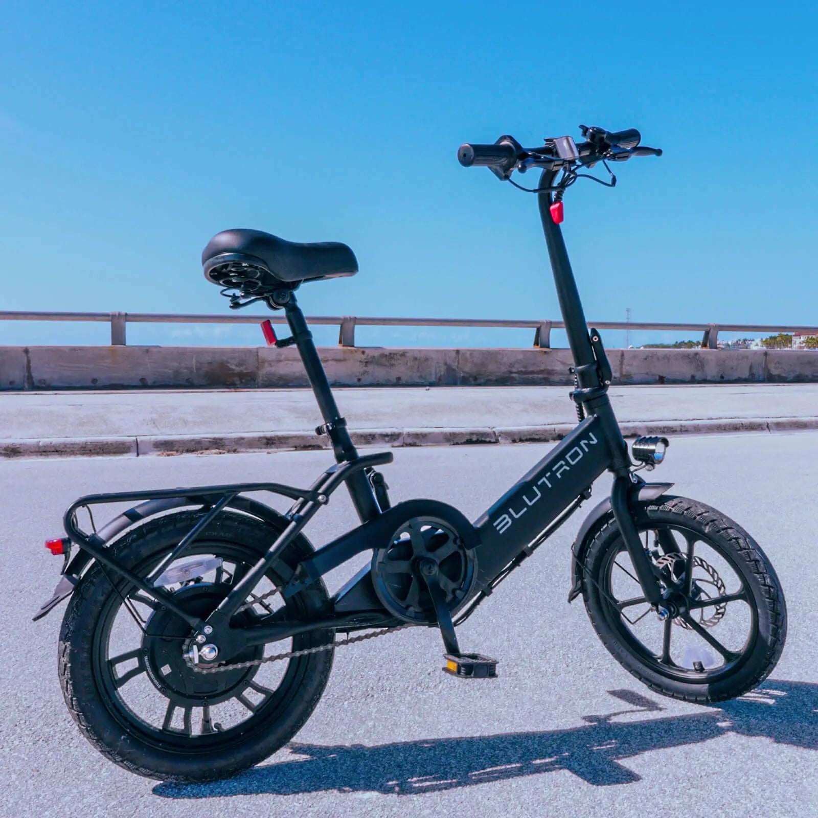 Blutron foldable ebike 350w (sleek black model) parked on paved outdoor ground: equipped with front LED light, rear cargo rack, adjustable padded seat, pedals, and front disc brake, set against a guardrail and clear blue sky.