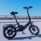 Blutron foldable ebike 350w (sleek black model) parked on paved outdoor ground: equipped with front LED light, rear cargo rack, adjustable padded seat, pedals, and front disc brake, set against a guardrail and clear blue sky.