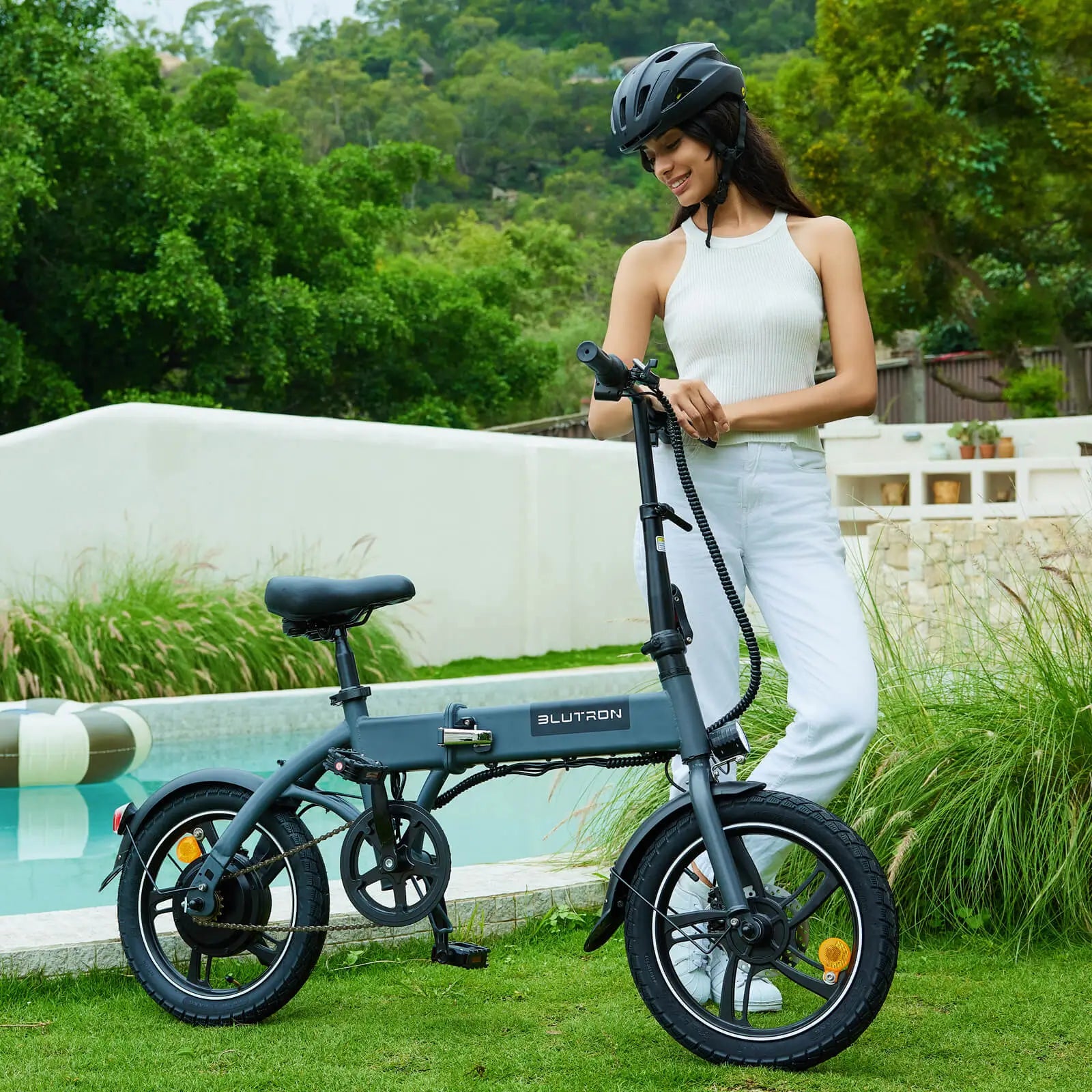 Helmeted woman standing by a BLUTRON folding ebike (among the best folding ebikes) on grass near a pool — the bike features a frame folding latch and a compact, portable design