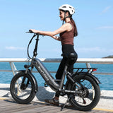 Woman (in a brown tank top, black leggings, and white helmet) on a coastal boardwalk, with a Blutron cheapest foldable ebike: This silver-gray model features "BLUTRON" and "EB750S" branding, a rear cargo rack, wide tires, front/rear disc brakes, set against the bright blue sea under a sunny sky.