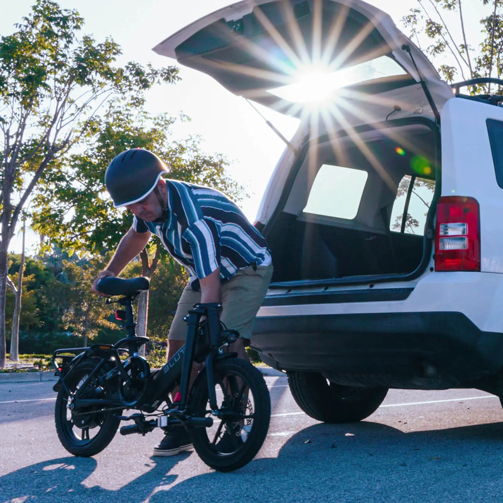 Helmeted rider (in a striped shirt and khaki shorts) loads a compact folding commuter ebike into the open trunk of a white SUV—sunlight flares behind, with leafy trees lining the outdoor parking area.