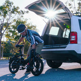 Helmeted rider (in a striped shirt and khaki shorts) loads a compact folding commuter ebike into the open trunk of a white SUV—sunlight flares behind, with leafy trees lining the outdoor parking area.