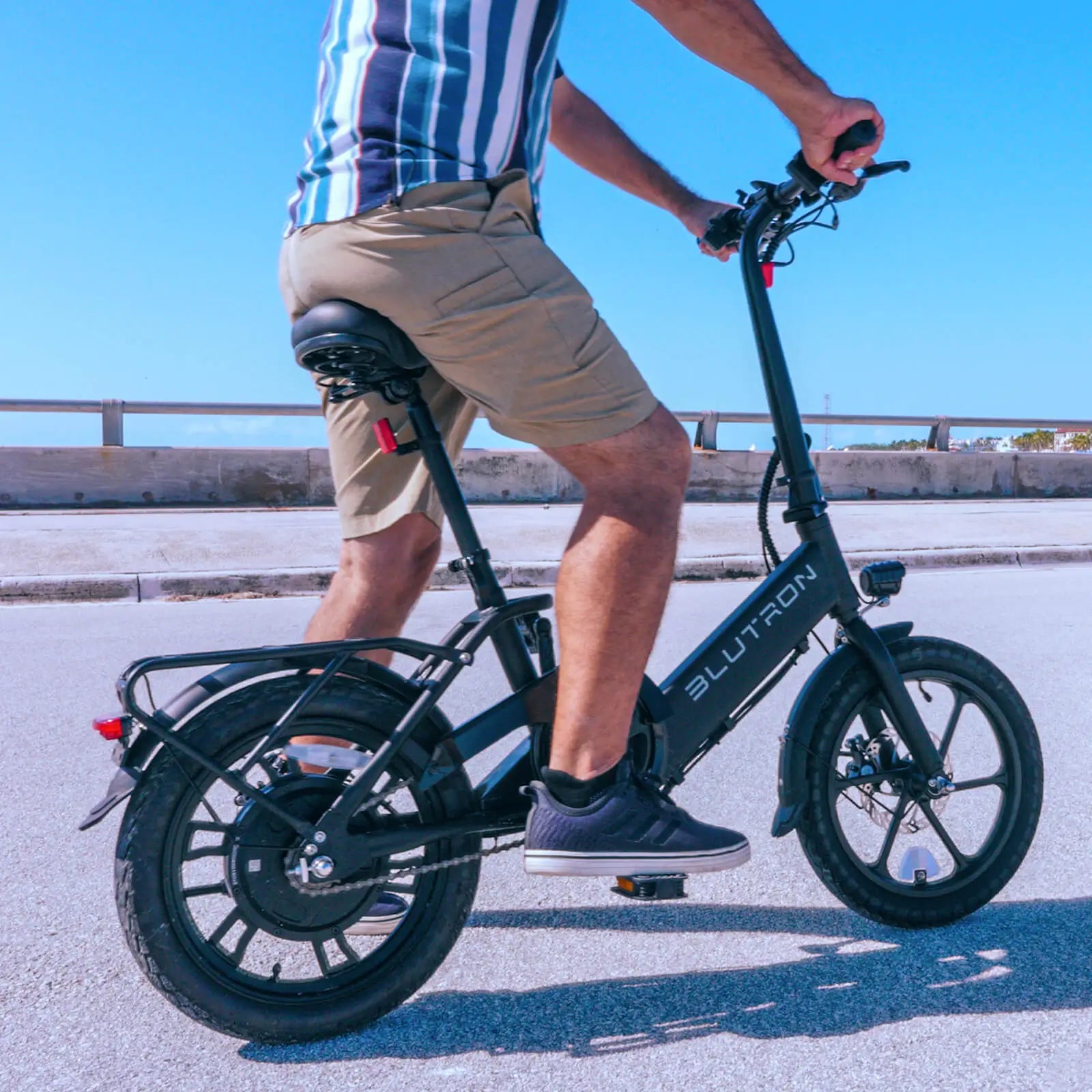 Rider pedals a sleek Blutron folding ebike along a paved roadside—fitted with rear cargo rack, taillight, padded seat, front LED light—against a bright sky and guardrail.