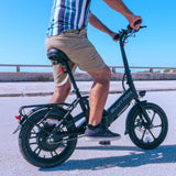 Rider pedals a sleek Blutron folding ebike along a paved roadside—fitted with rear cargo rack, taillight, padded seat, front LED light—against a bright sky and guardrail.