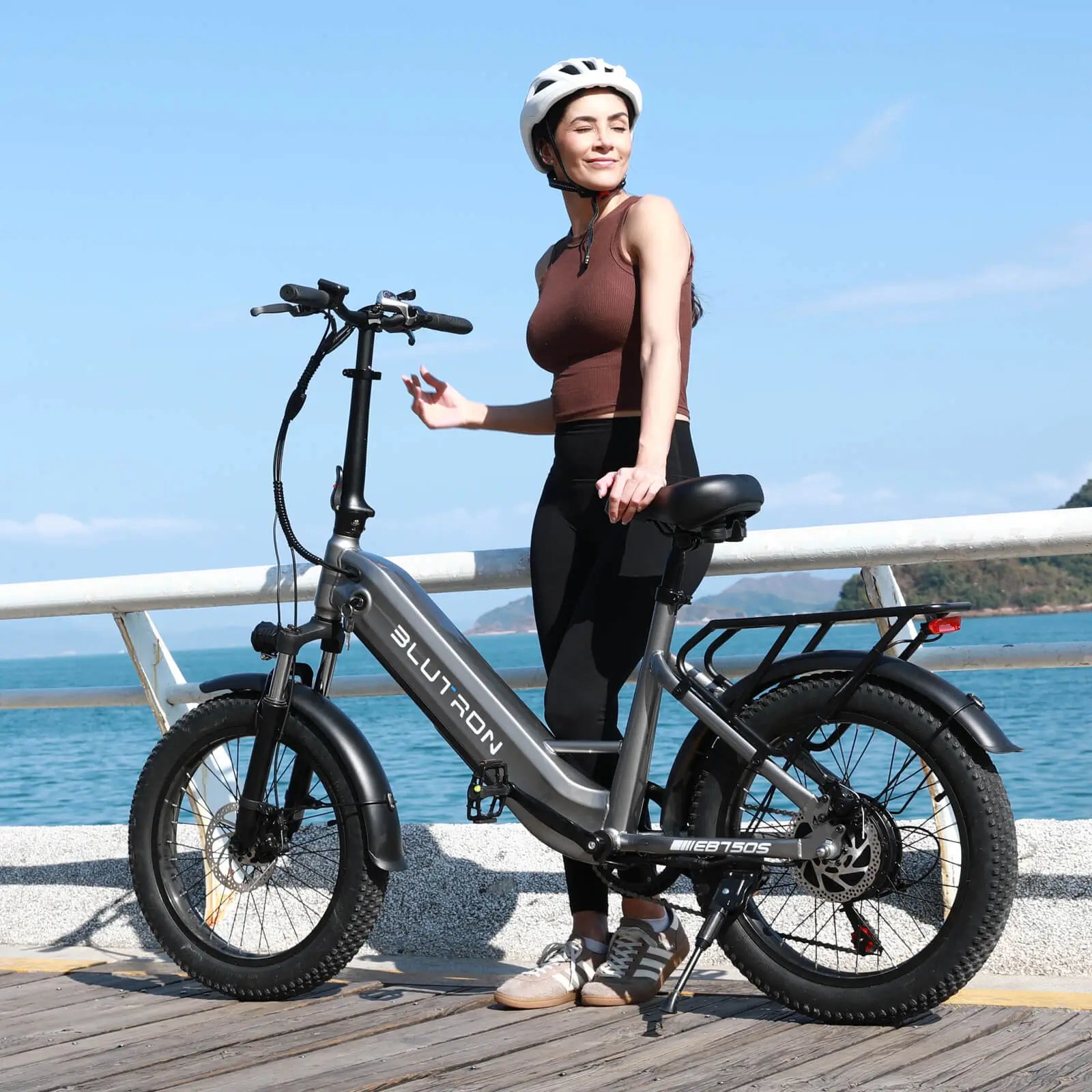 Woman (brown sleeveless top, black leggings, white helmet) by a Blutron portable folding electric bike for urban leisure (EB750S) on a coastal boardwalk: Silver-gray model with "BLUTRON" branding, rear rack, wide tires, disc brakes; backdrop of blue sea & sunny sky.