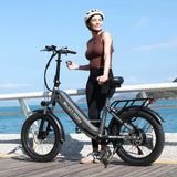 Woman (brown sleeveless top, black leggings, white helmet) by a Blutron portable folding electric bike for urban leisure (EB750S) on a coastal boardwalk: Silver-gray model with "BLUTRON" branding, rear rack, wide tires, disc brakes; backdrop of blue sea & sunny sky.