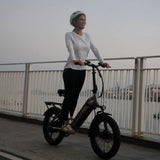 Woman (white top, black leggings, helmet): Riding a Blutron compact folding e-bike for urban evening rides (branded) on a dusk waterfront promenade; has rear rack, wide tires; port cranes, calm waters backdrop