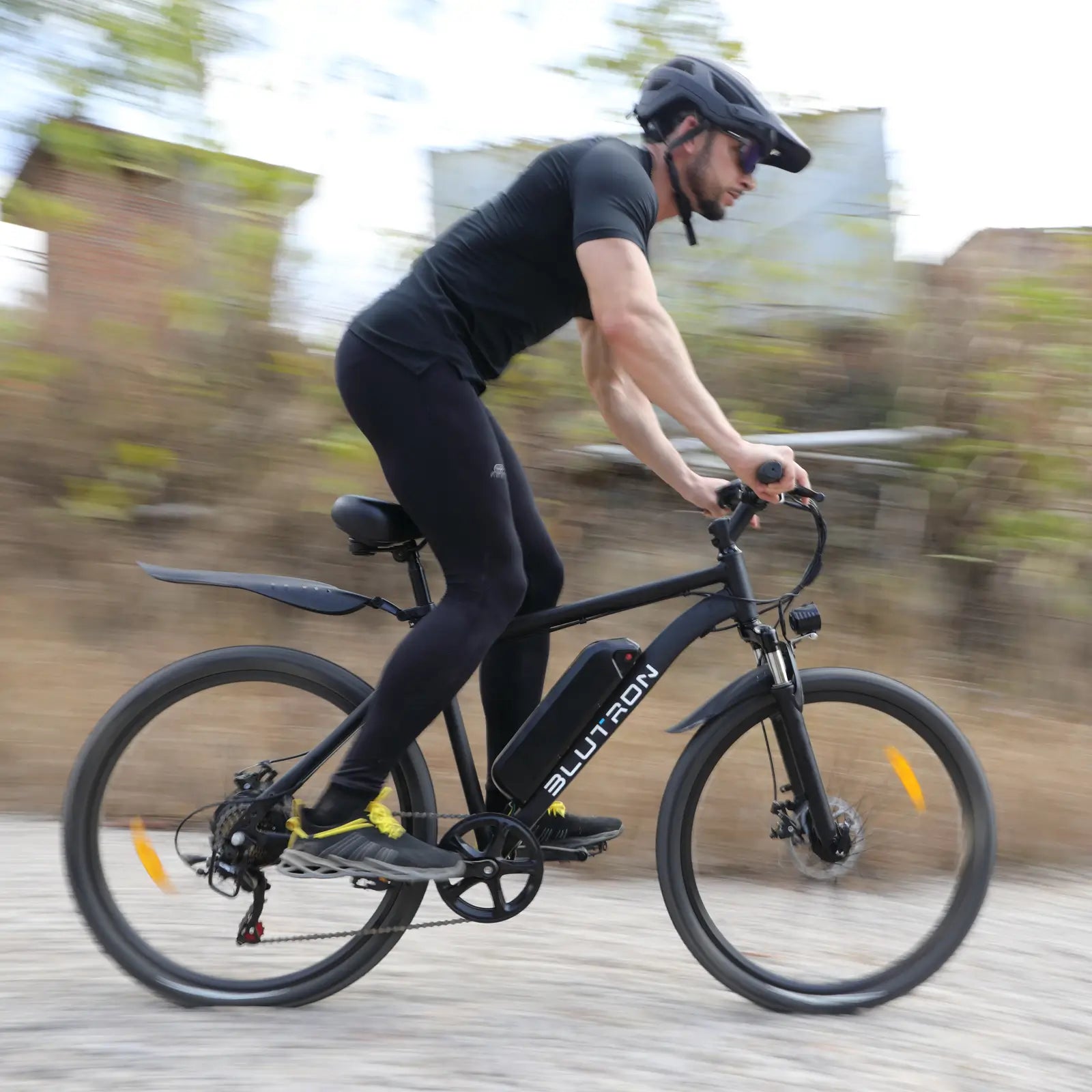 BLUTRON EB550 electric mountain bike: Rider in athletic gear cycling dynamically on a gravel path, showcasing the bike’s detachable BLUTRON-branded battery — set against an outdoor rural backdrop