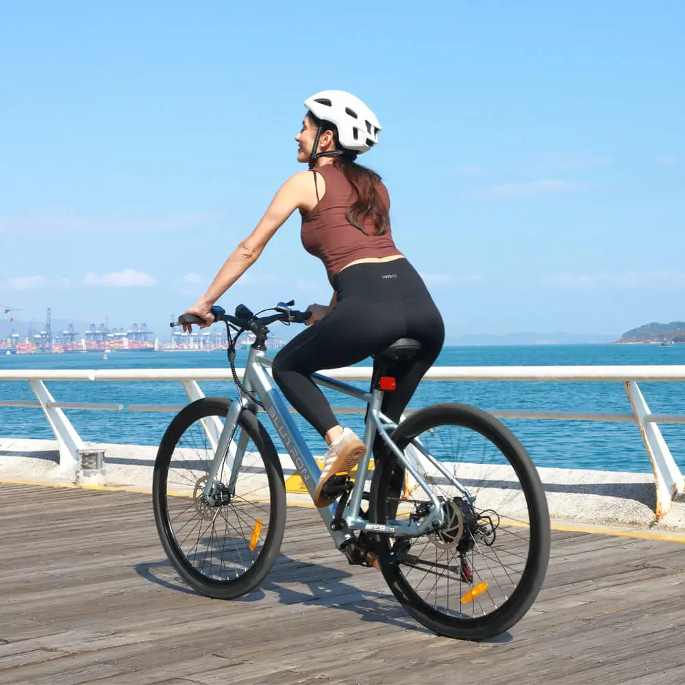 Helmeted woman rides the Blutron EB570 lightweight commuter ebike along a coastal boardwalk. Its portable build and smooth frame suit daily commutes and scenic rides (sea, port backdrop).