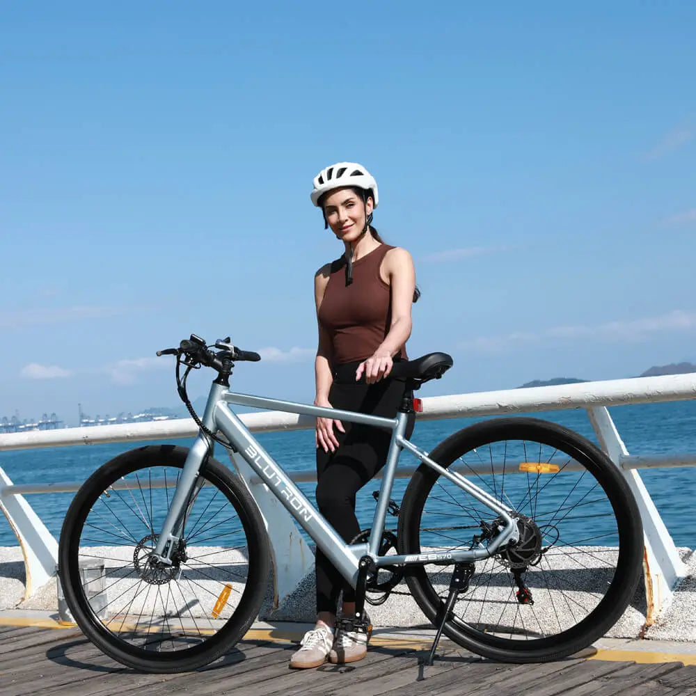 Helmeted woman stands by the Blutron EB570 long range commuter ebike on a coastal boardwalk. Its extended battery life fits daily commutes and scenic coastal rides (sea, distant landscapes backdrop).