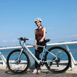 Helmeted woman stands by the Blutron EB570 long range commuter ebike on a coastal boardwalk. Its extended battery life fits daily commutes and scenic coastal rides (sea, distant landscapes backdrop).